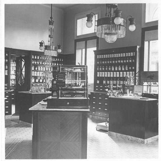 Historische Aufnahme der Offizin mit Holzregalen, Standgefäßen, Kronleuchtern und einer alten Registrierkasse